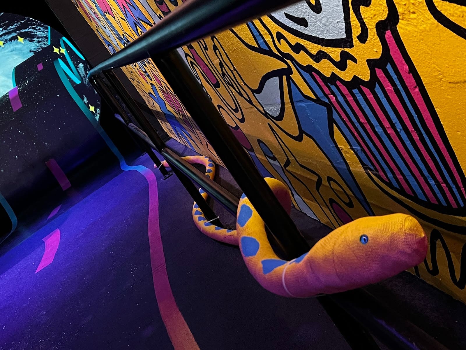 Colorful stuffed snake with blue spots on a handrail in a vibrant, graffiti-style room with bright yellow and blue patterns.
