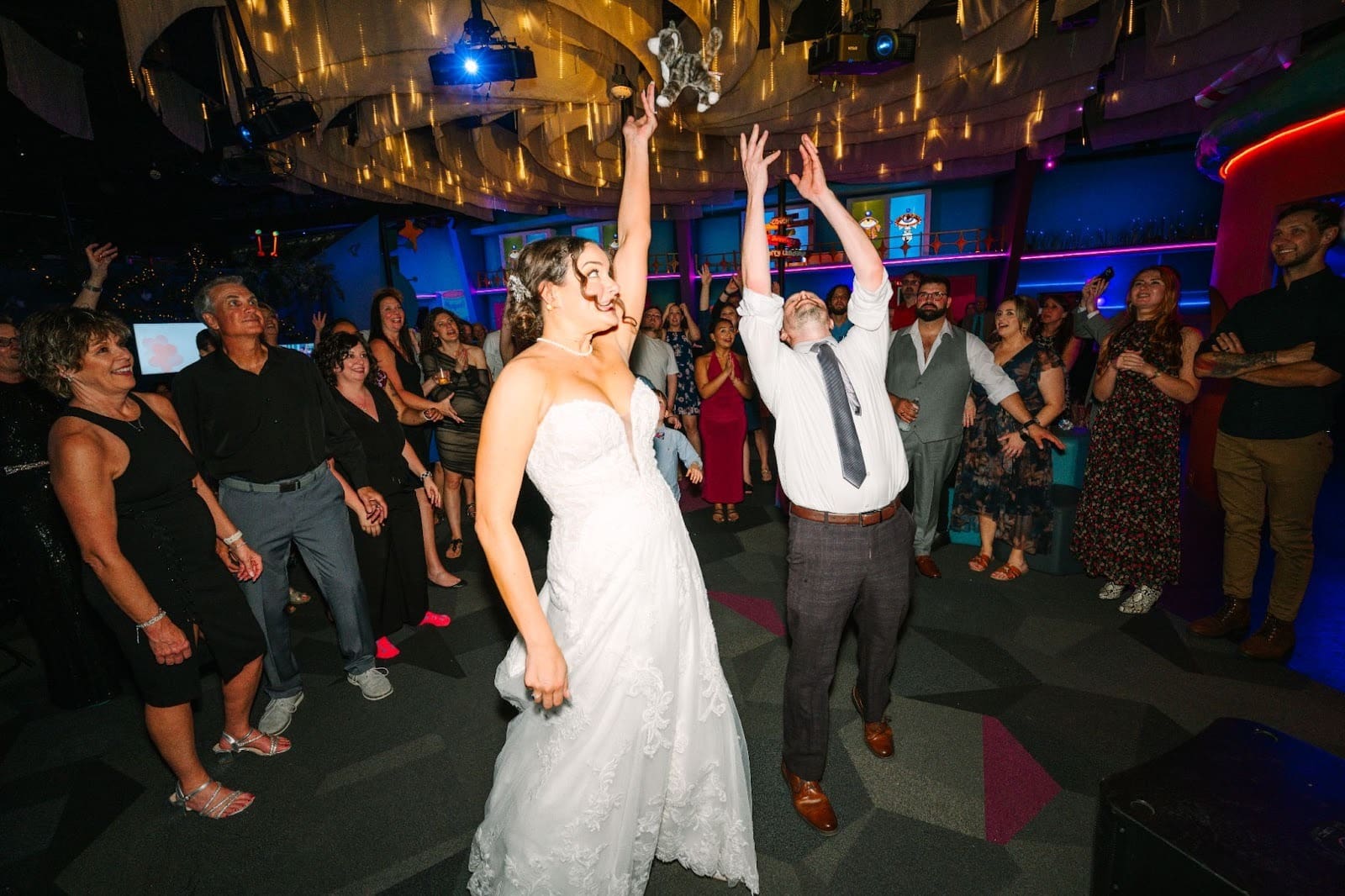 Bride in white dress throws bouquet, man in white shirt reaches up. Guests cheer in a vibrant, decorated hall with colorful lights an example of Unique Wedding Ideas