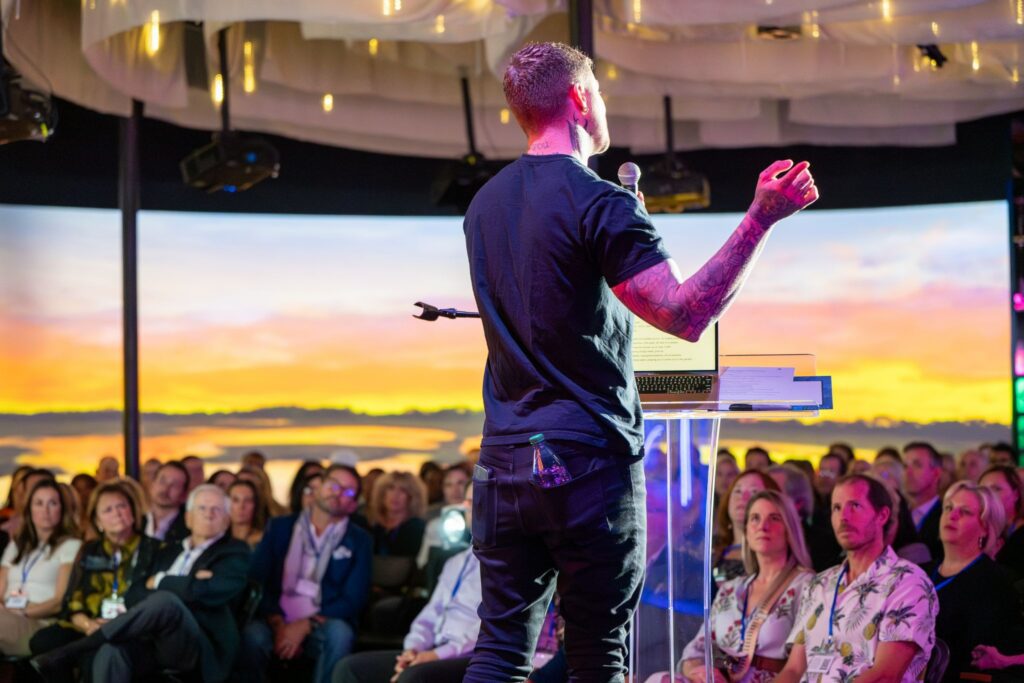 A speaker addresses a seated audience in an immersive event space featuring a large digital sunset backdrop
