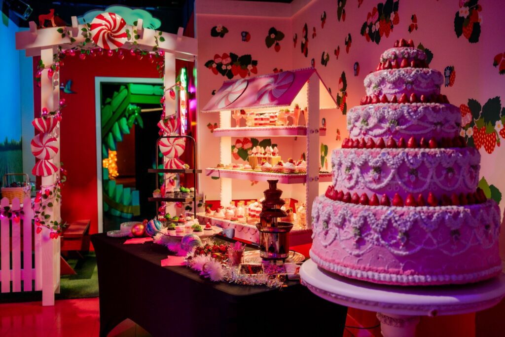 A whimsical dessert table with a large tiered pink cake and chocolate fountain in a strawberry-themed immersive art room