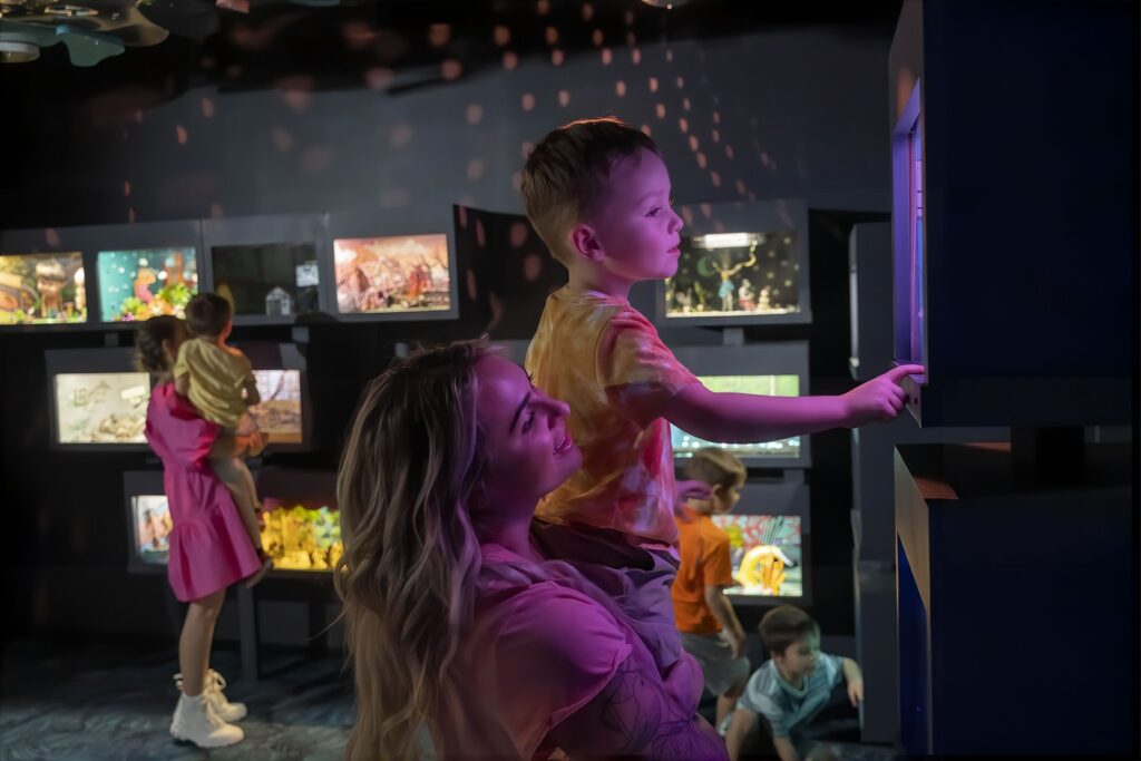 A woman holds a young boy as he interacts with a glowing display in a room filled with illuminated dioramas