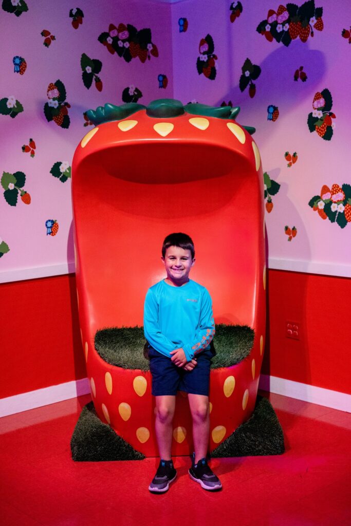 A boy smiles while sitting inside a giant red strawberry-shaped chair in an immersive art exhibit