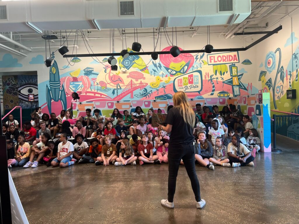 A guide leads a large group of students sitting on the floor in front of a vibrant, Florida-themed mural