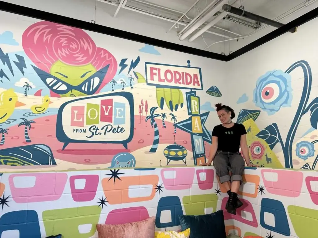 Ashley Cantero sits in front of a colorful retro mural featuring the text Love from St. Pete and FloridaRama